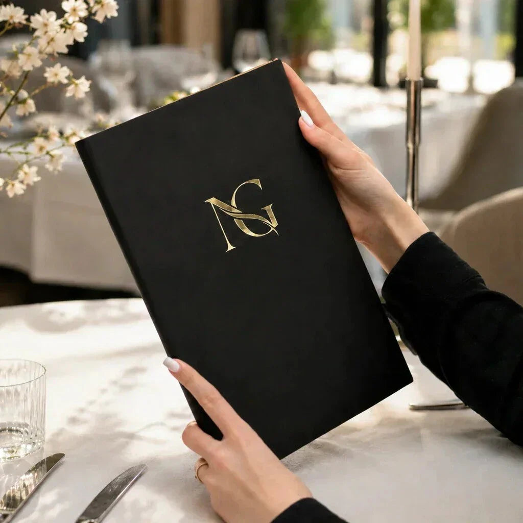 Black leather menu cover with gold logo in a bright restaurant setting with white tablecloth, glassware, flowers, and soft natural light.