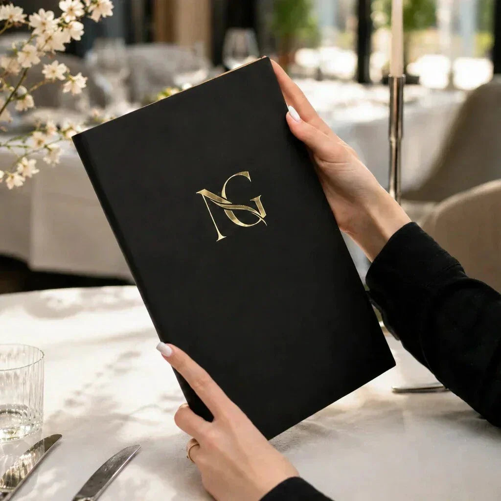 Black leather menu cover with gold logo in a bright restaurant setting with white tablecloth, glassware, flowers, and soft natural light.
