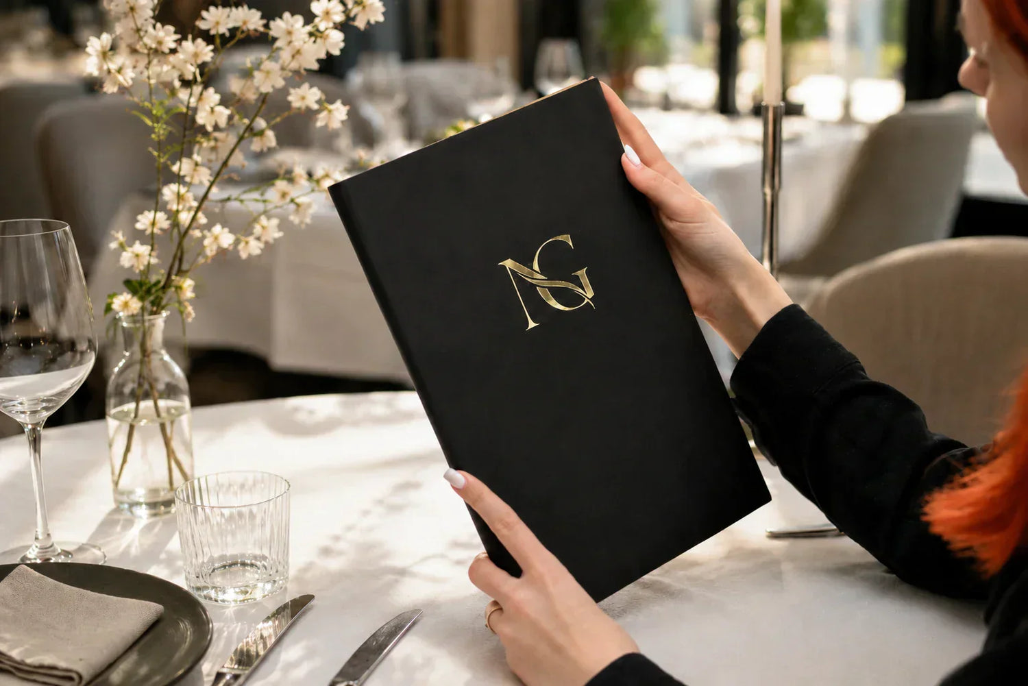 Black leather menu cover with gold logo in a bright restaurant setting with white tablecloth, glassware, flowers, and soft natural light.