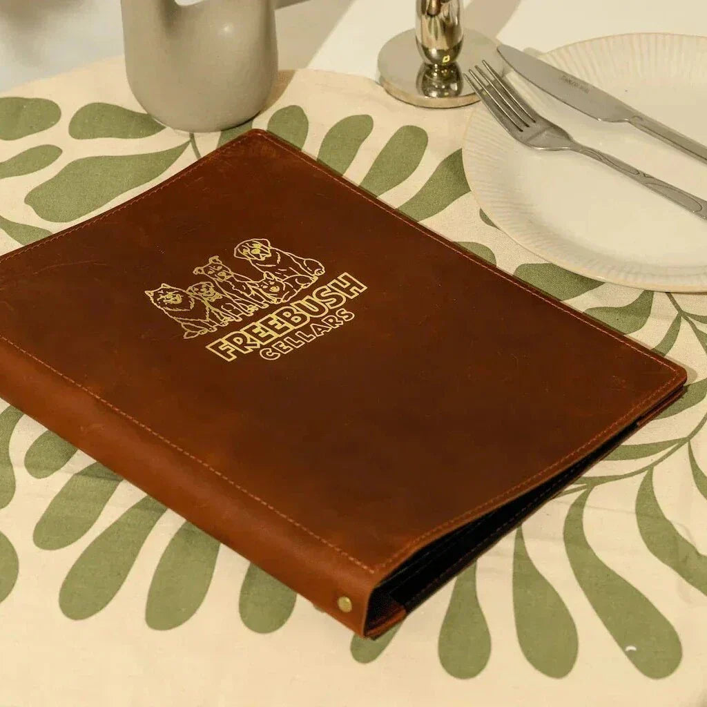 Handcrafted brown leather menu folder with custom gold foil branding and professional bolt binding, shown in a premium table setting