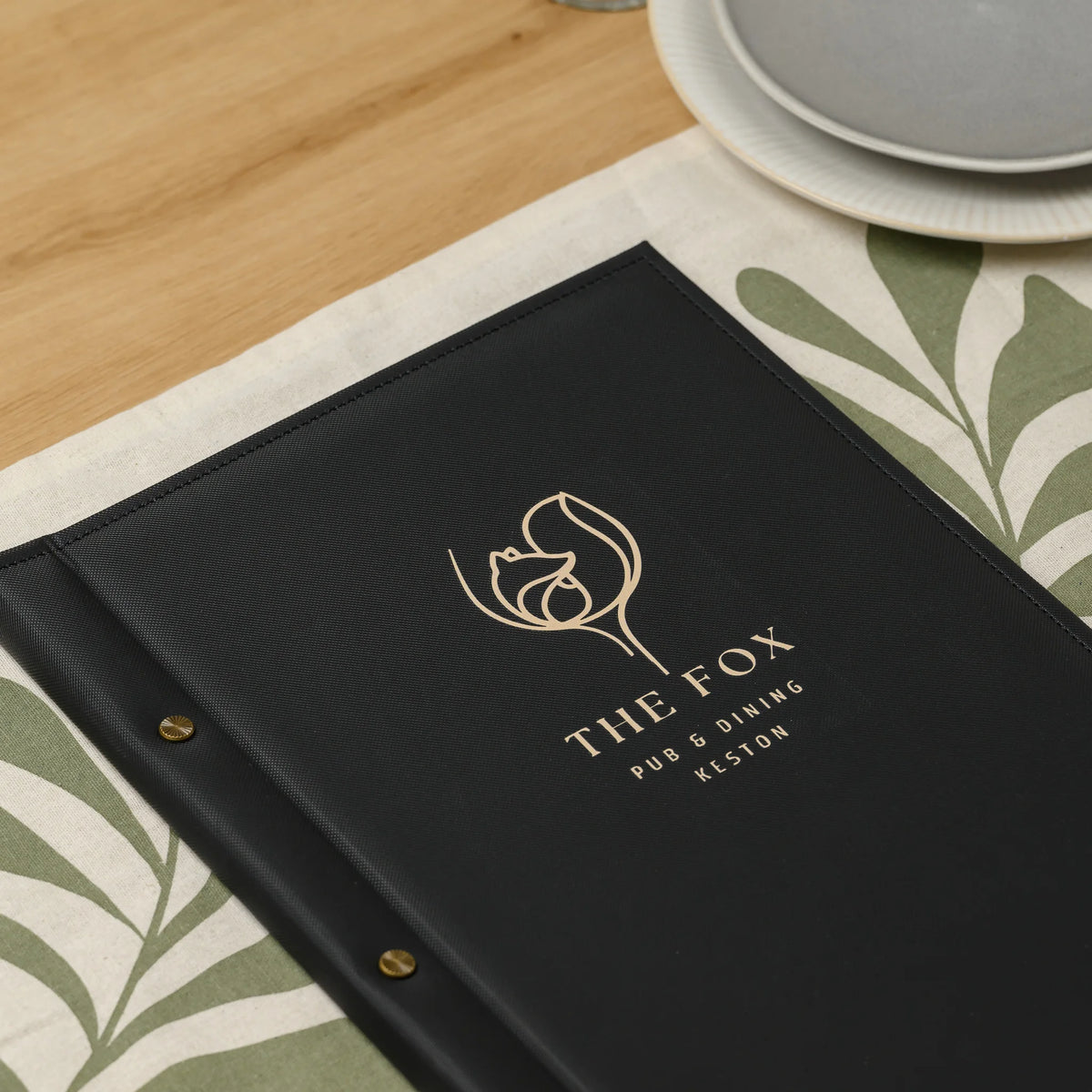 Black faux leather menu cover with a gold printed logo and three brass screw heads lying on a set dining table