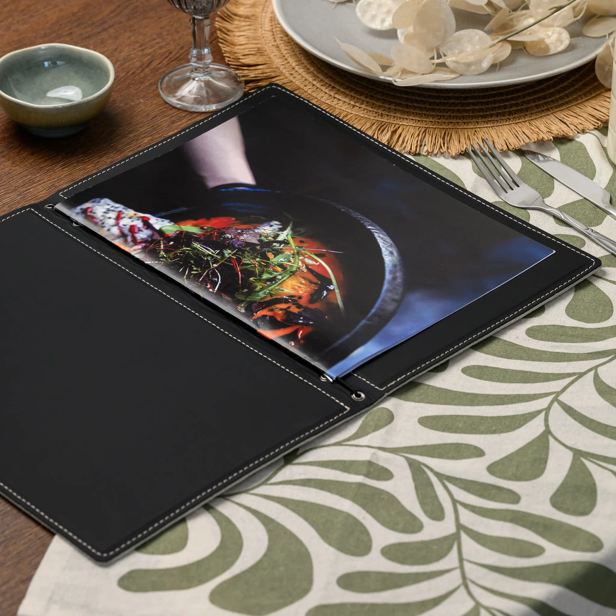 Open menu folder lying on a set table with a food photo insert, black lining and silver eyelets along the spine