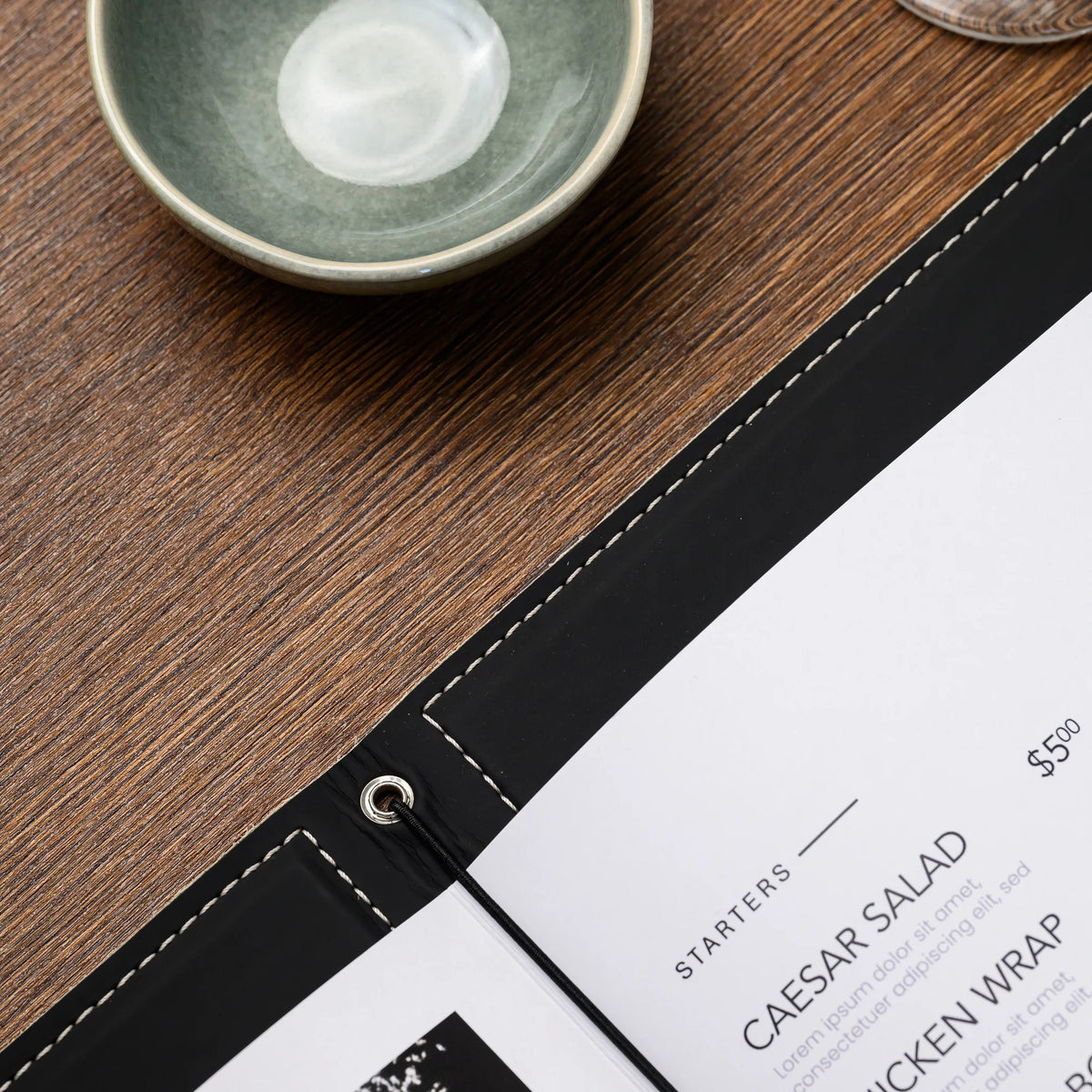 Top view of a silver eyelet with black elastic cord holding printed menu pages, ceramic bowl on wooden table