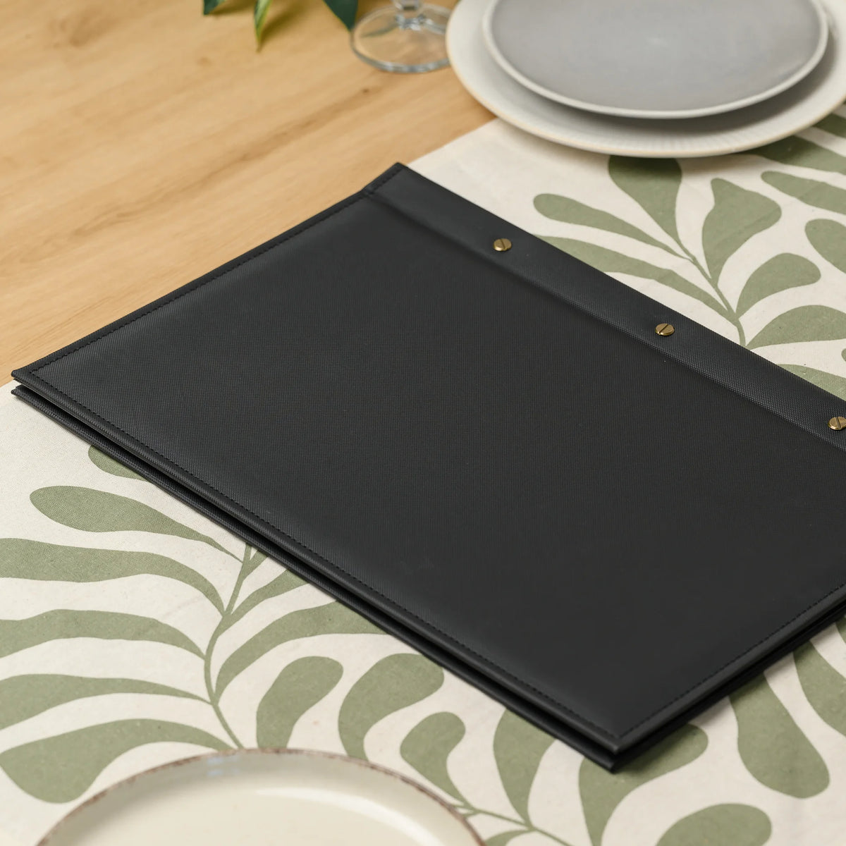 Back of a black textured faux leather menu cover with three slotted brass screws lying on a patterned table runner