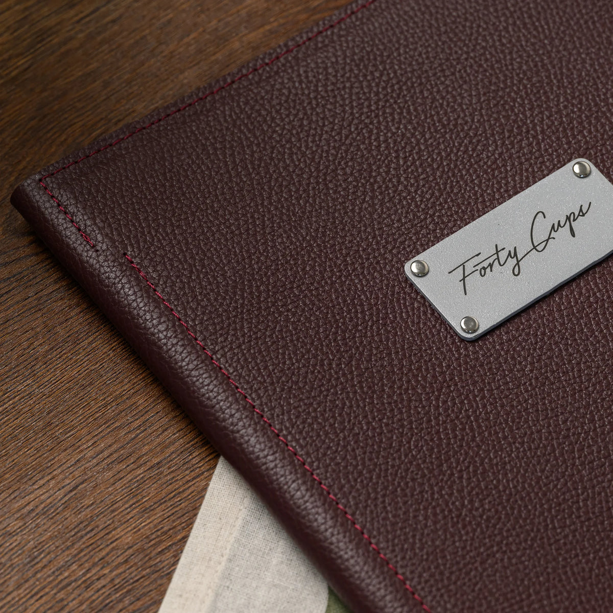 Close-up of bordeaux pebble-grain leather cover with red stitching and a riveted silver logo plate on wood