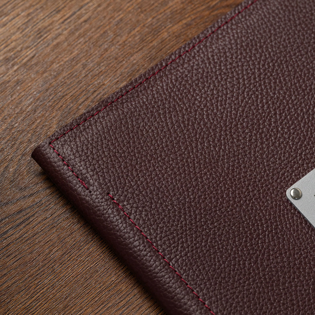Corner of bordeaux leather menu cover with red edge stitching and the edge of a silver plate on a wooden table