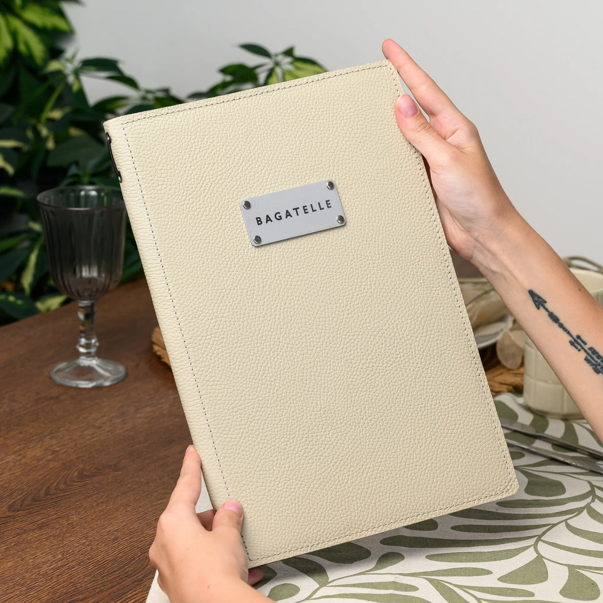 Hands holding a closed beige leather menu folder upright with a rectangular silver logo plate near the top