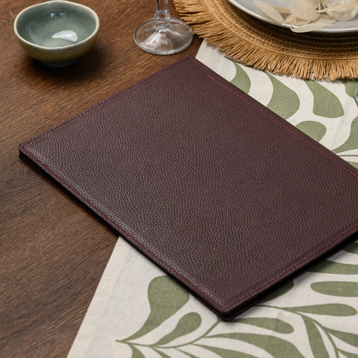 Wide view of plain bordeaux leather menu cover lying on a table runner beside a ceramic bowl and wine glass