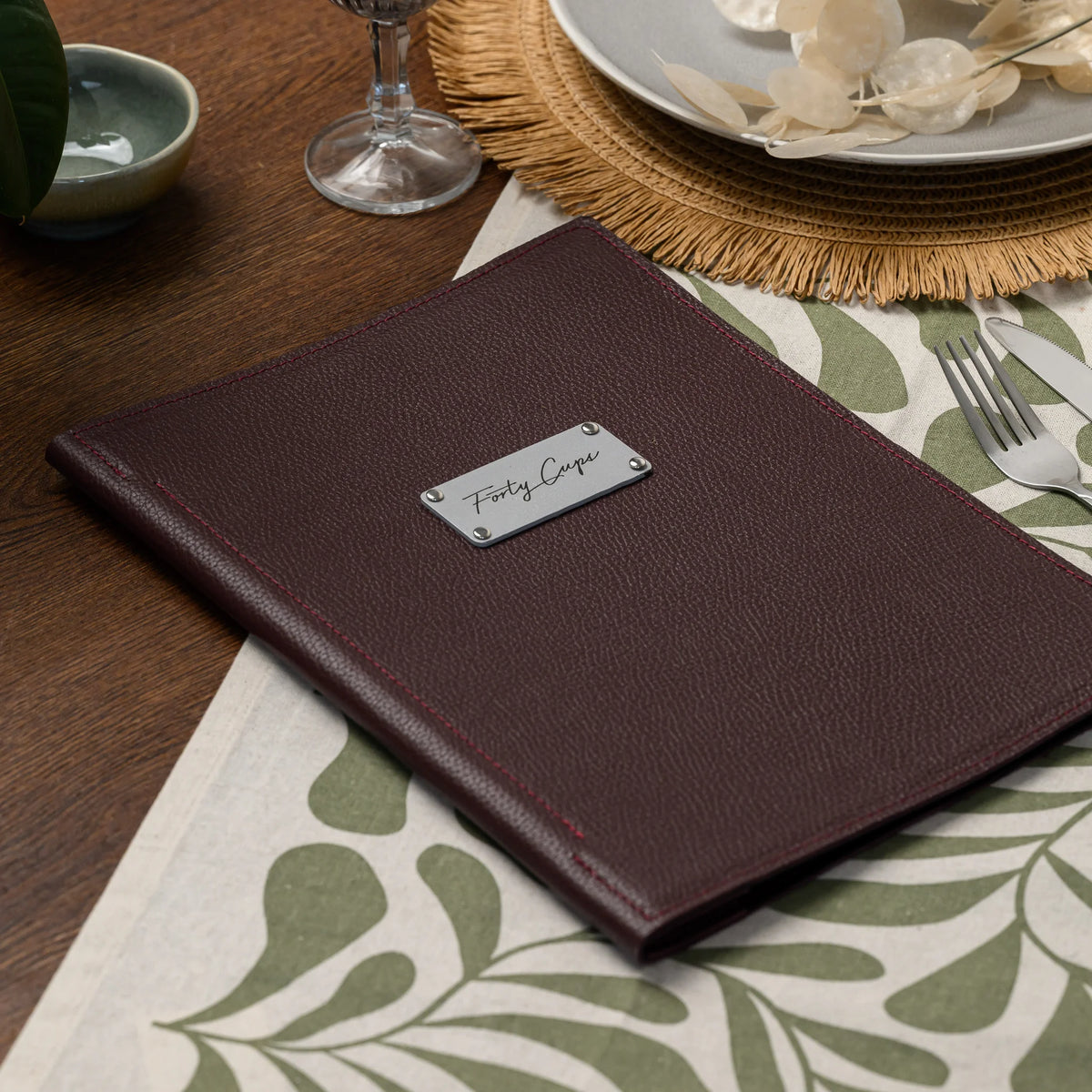 Closed bordeaux leather menu cover with a rectangular silver logo plate on a set table next to cutlery
