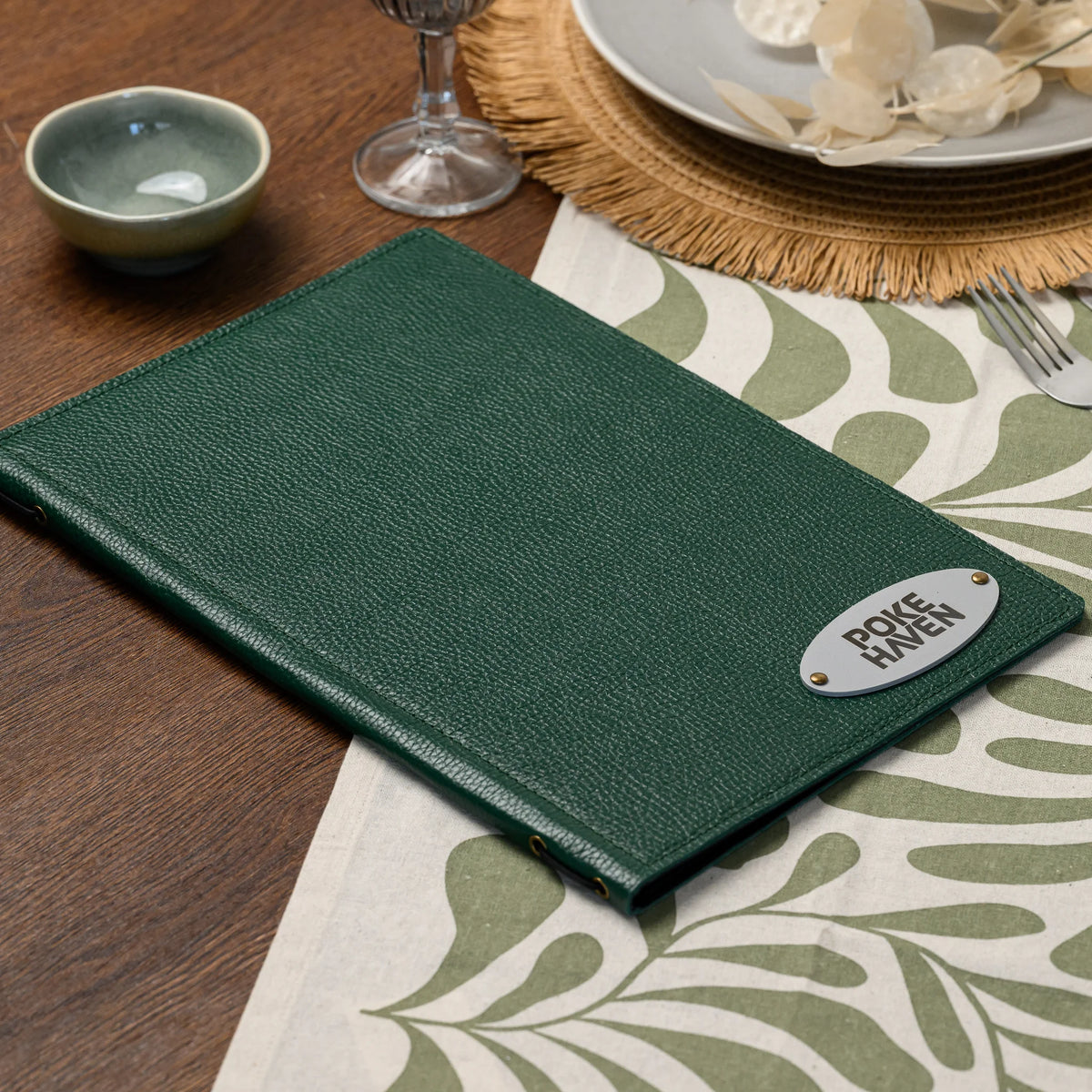 Closed green leather menu folder with oval silver logo plate on a set table beside a ceramic bowl and wine glass