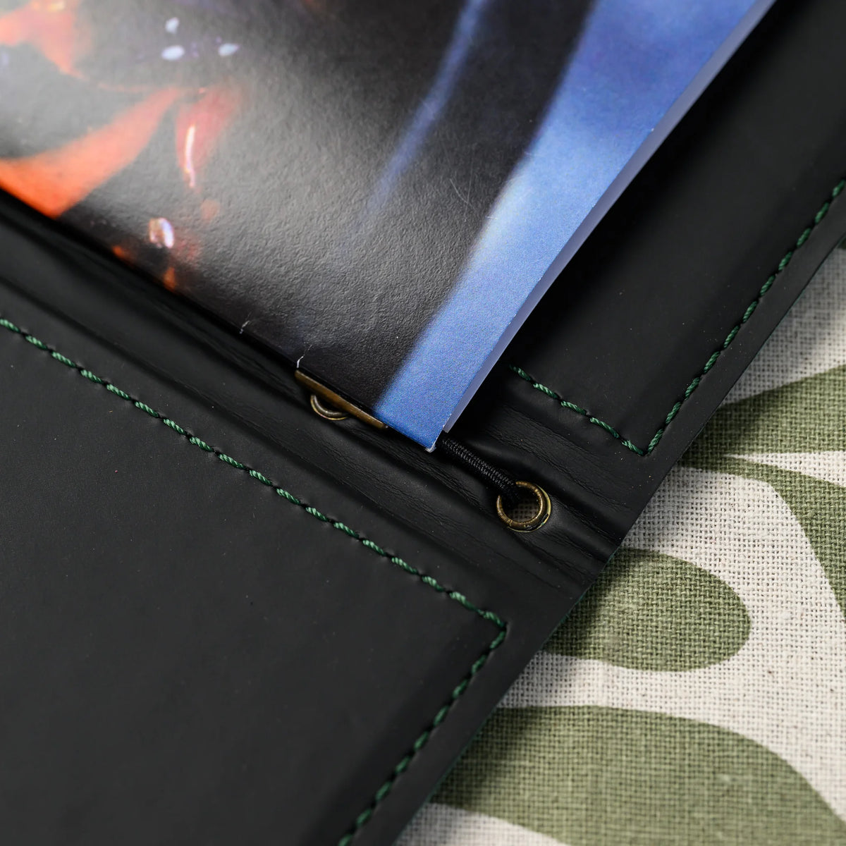 Macro of folder spine interior with black elastic cord, brass eyelet and small brass stopper holding folded pages