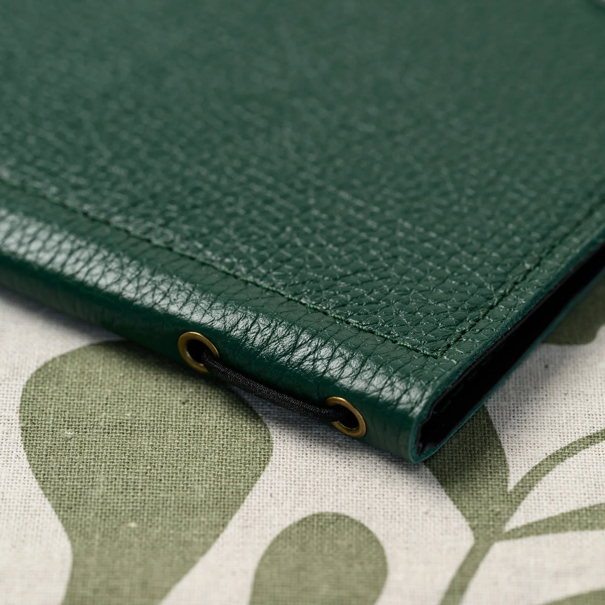 Detail of green leather menu folder spine with two brass eyelets and black elastic band on light linen cloth