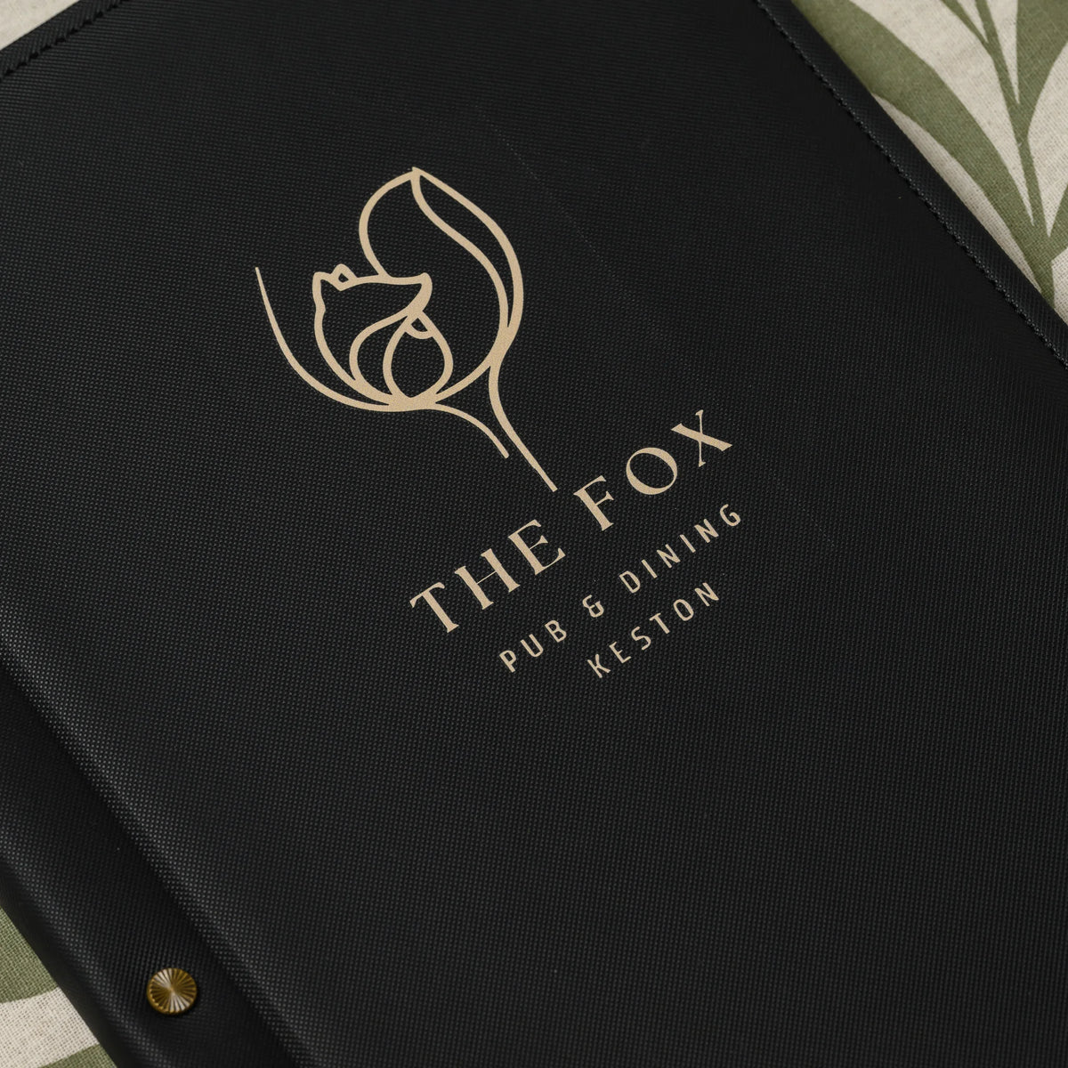 Close-up of a gold printed logo with fine line details on the front of a black textured faux leather menu cover