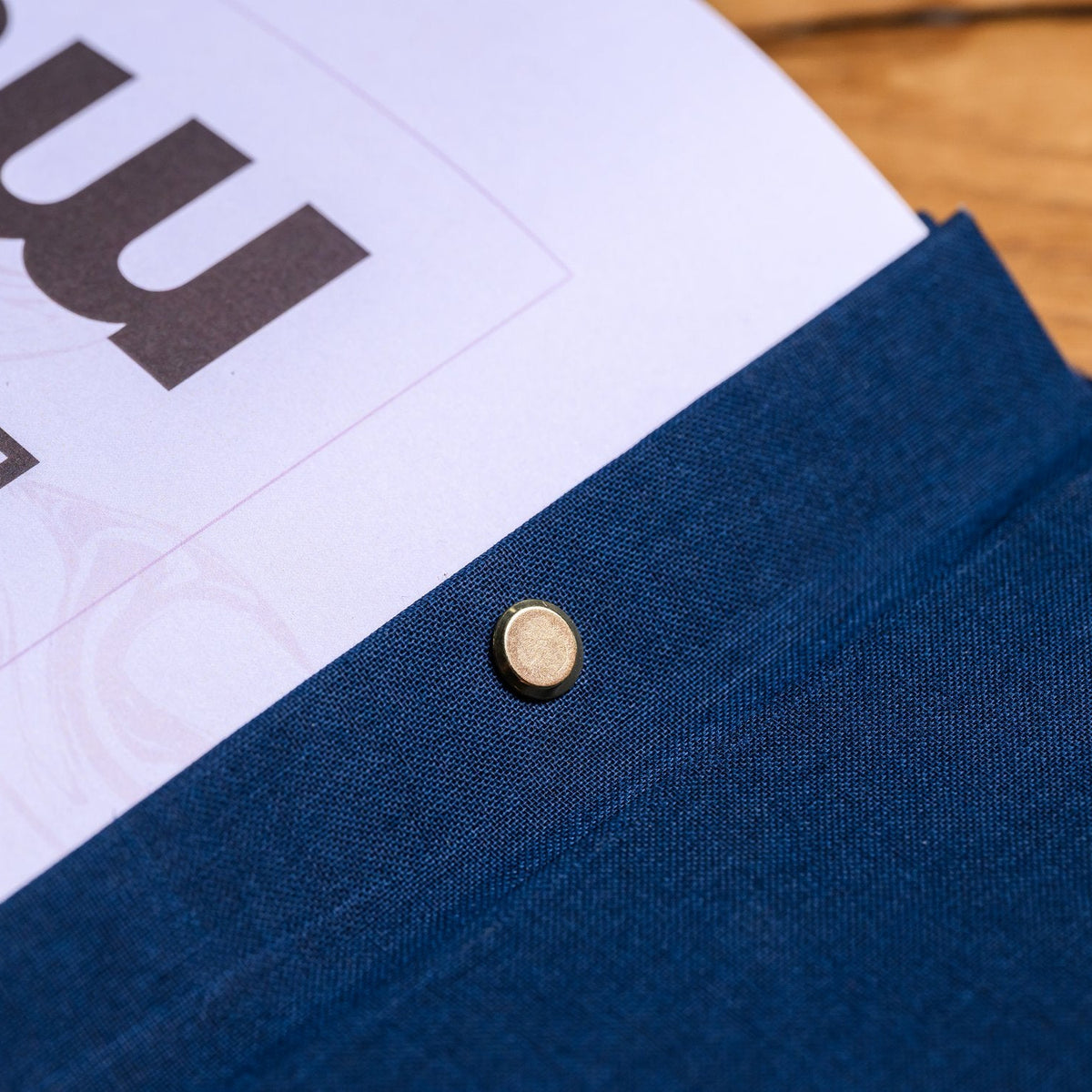 Close-up of a closed navy textile folder corner with a gold snap fastener against a dark blurred background