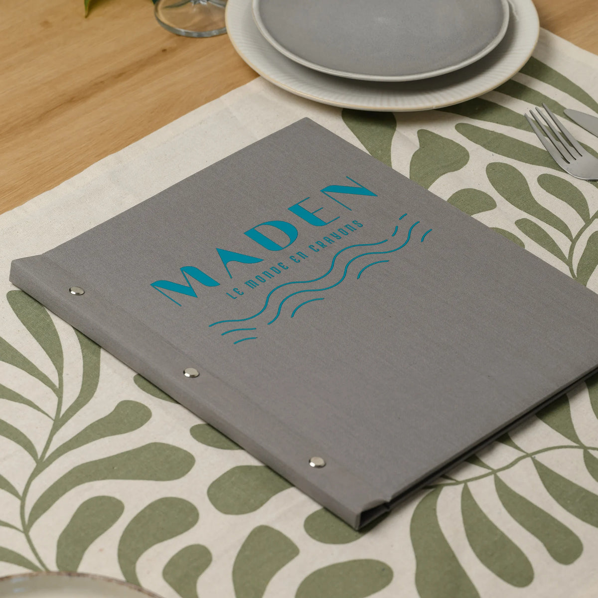 Gray fabric menu cover with a full-color DTF printed logo and three chrome screws lying on a set dining table