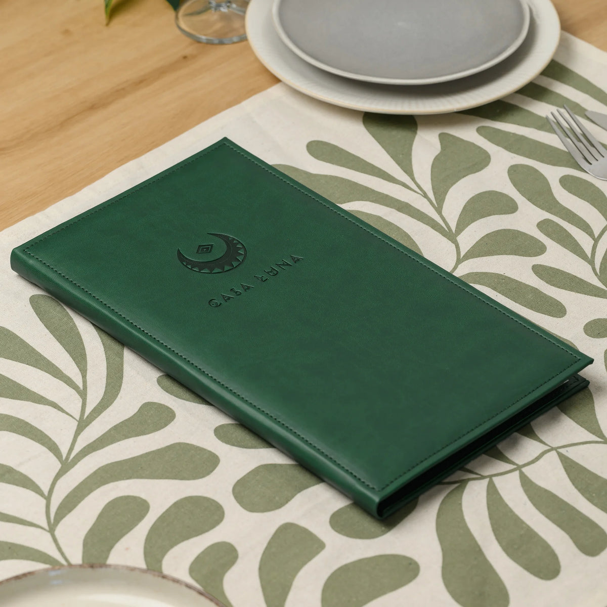 Closed green faux leather menu cover with a blind embossed logo lying on a patterned table runner next to plates