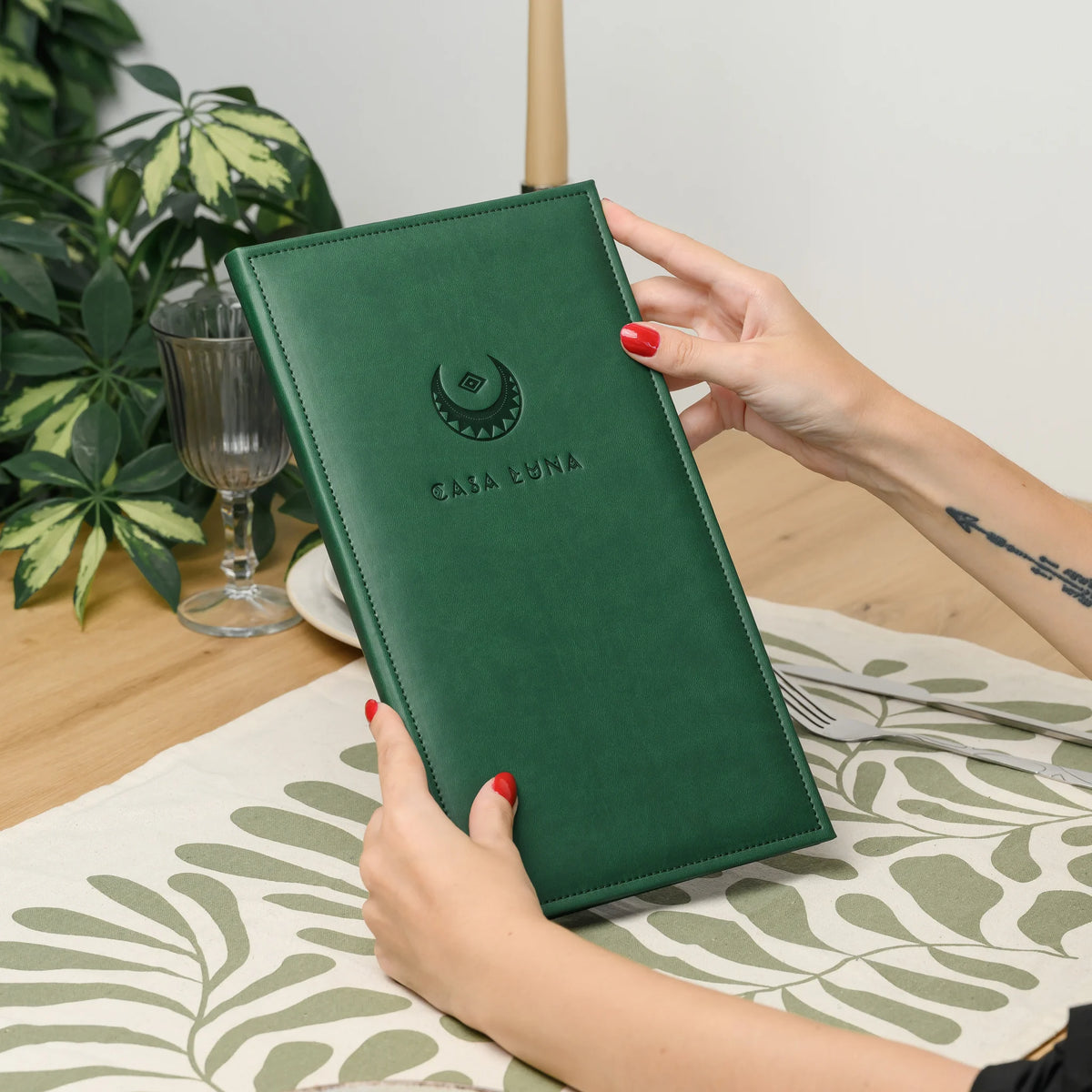 Hands holding a closed green faux leather menu cover with a blind embossed logo at a table with a glass and plant