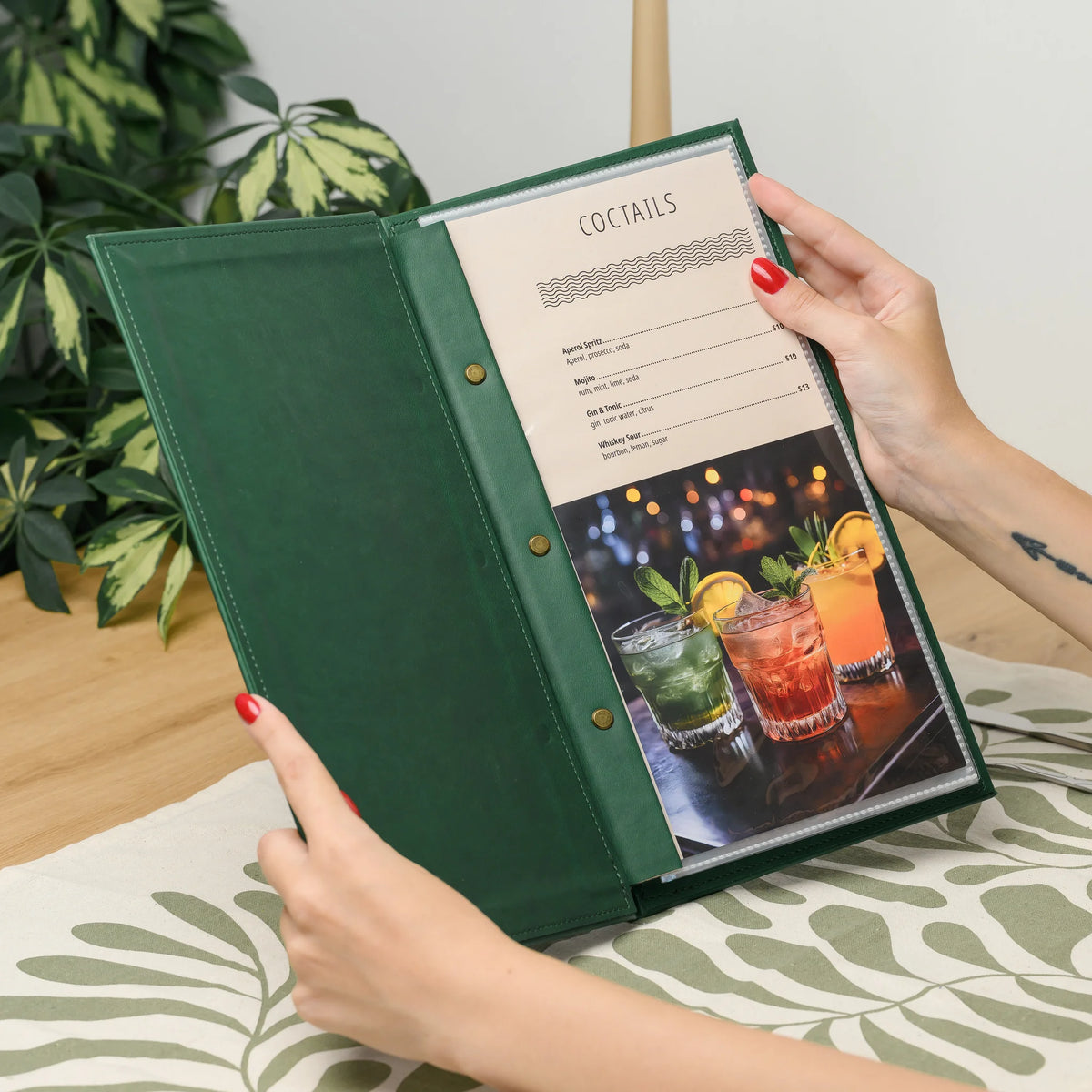 Hands holding an open green faux leather menu cover with a cocktail menu page in a clear sleeve and brass screws