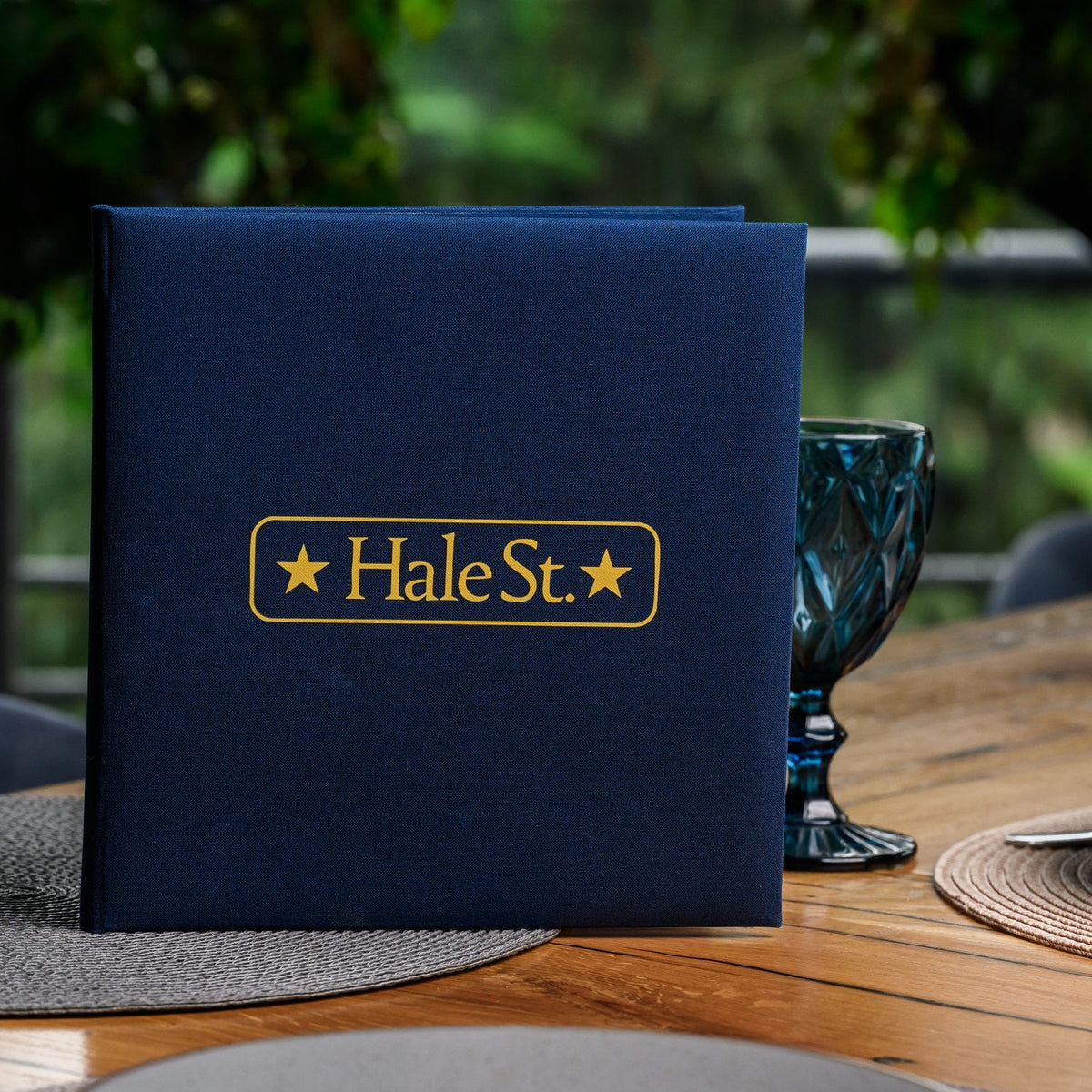 Closed navy textile menu folder standing upright with a gold star-badge logo against green outdoor foliage