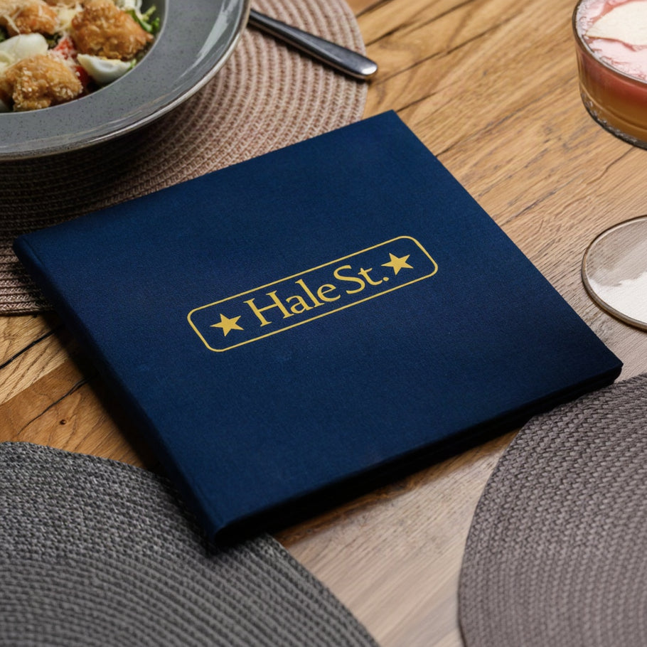 Small closed navy textile menu folder standing with a gold star-badge logo on a grey woven table placemat