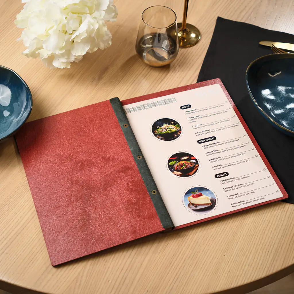 Open chili red plywood menu folder displayed on a round wooden table in a restaurant setting. White flowers and a dark blue ceramic bowl are visible in the background alongside golden candlesticks. A printed menu insert with food photos is visible inside. The deep red plywood cover and black elastic band create a bold and elegant table presentation for modern restaurants and upscale dining venues.