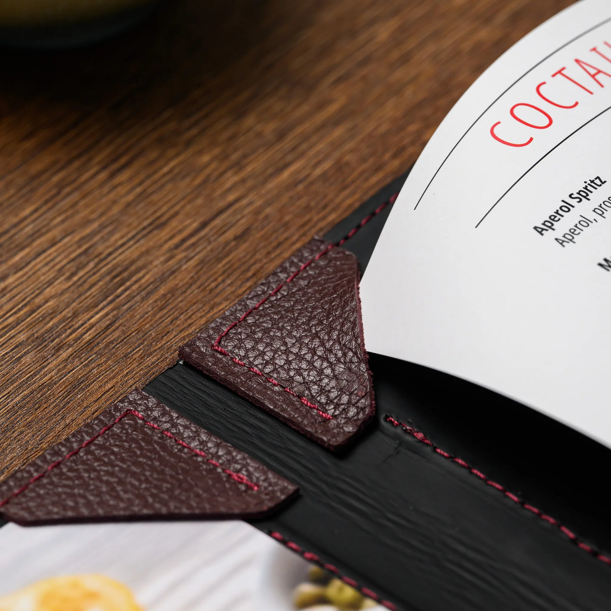 Close-up of bordeaux leather corner tabs holding a curved printed menu sheet at the spine of an open cover
