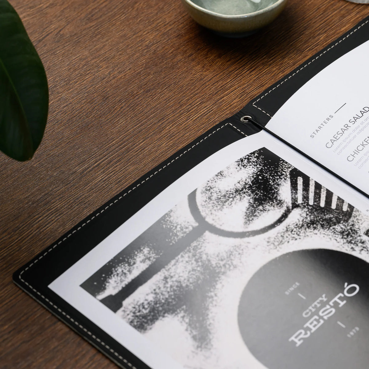 Open menu folder on wooden table with printed pages held by black elastic cord through a silver eyelet at the spine