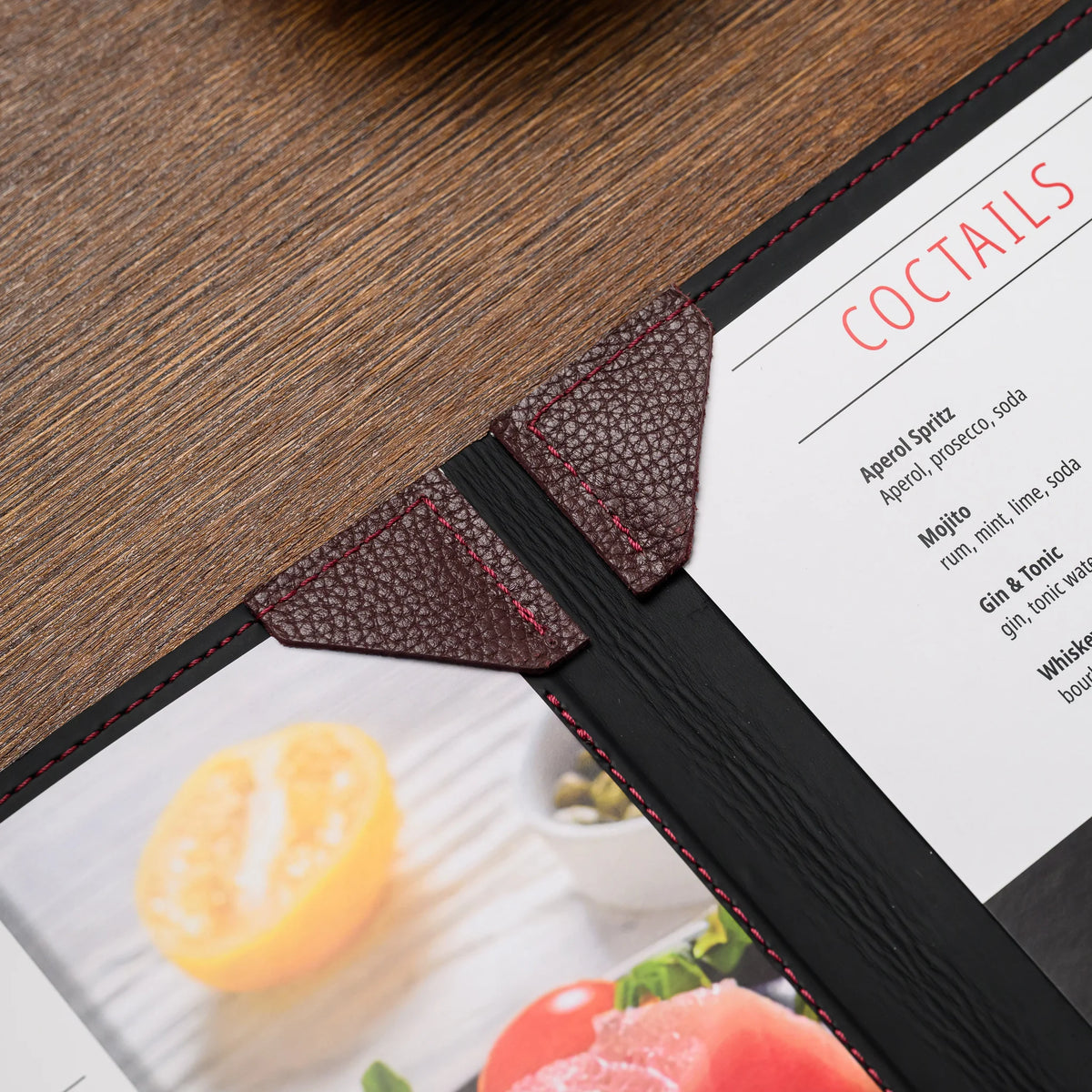 Close-up of bordeaux leather corner mountings with red stitching holding menu sheets at the spine on a wood table