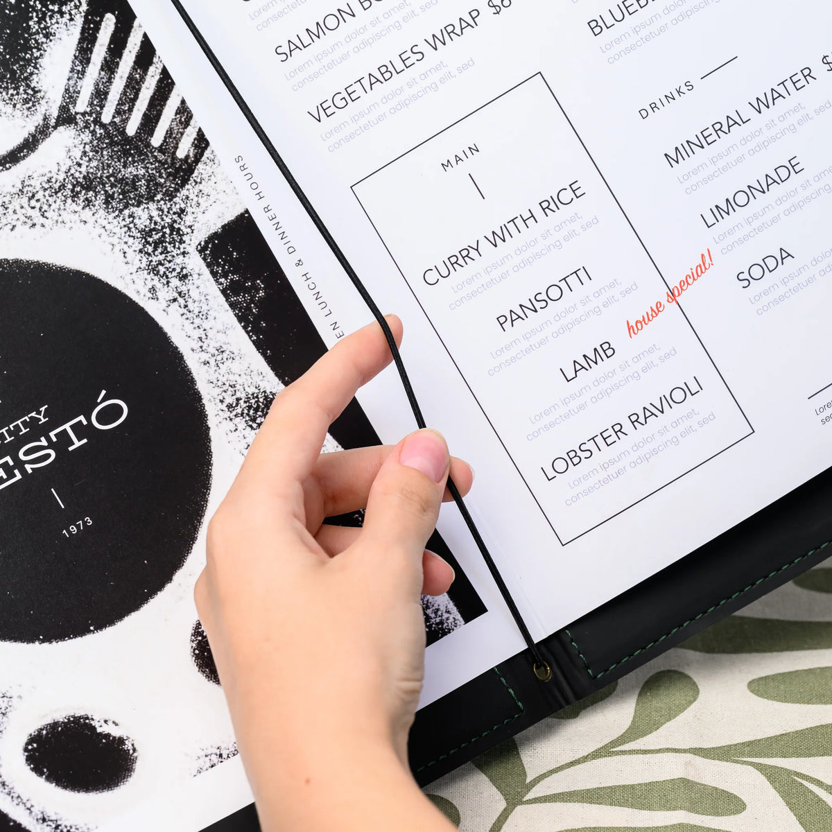 Hand pulling black elastic cord over printed menu pages inside an open folder, brass eyelet set into the spine
