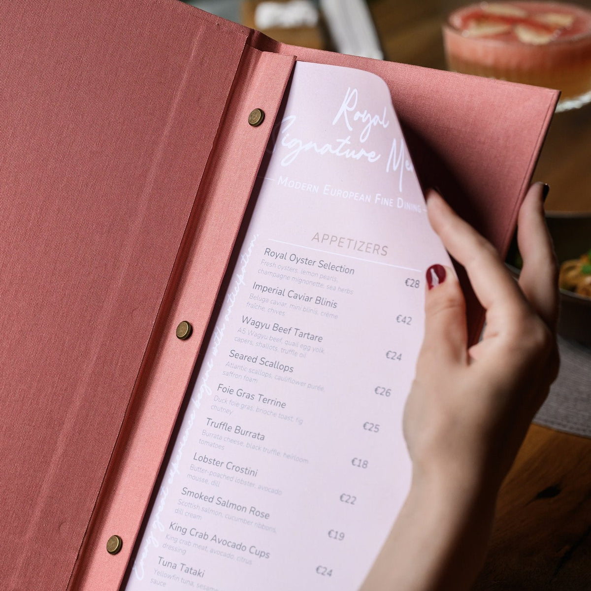 Hand holds a wide open coral-pink textile menu folder with a lavender printed menu page on wooden table