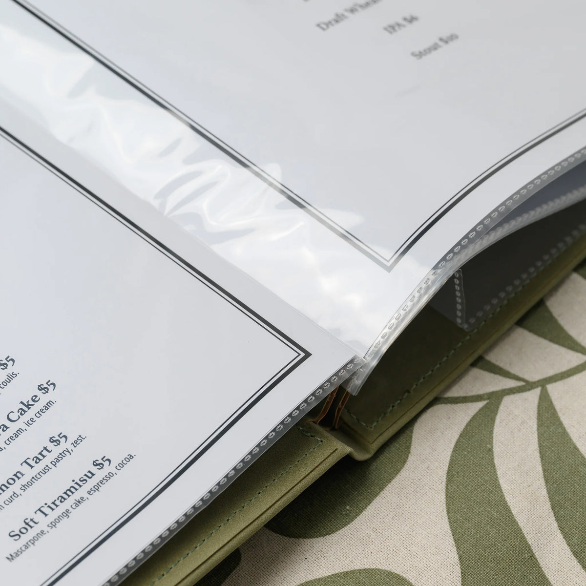 Close-up of a two-page menu spread in clear plastic sleeves turning at the spine of an olive faux leather cover