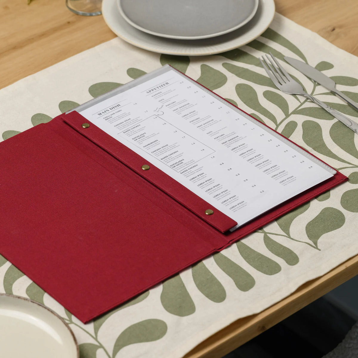 Open red fabric menu cover on a set table with a printed menu in a clear sleeve held by a plank with brass screws
