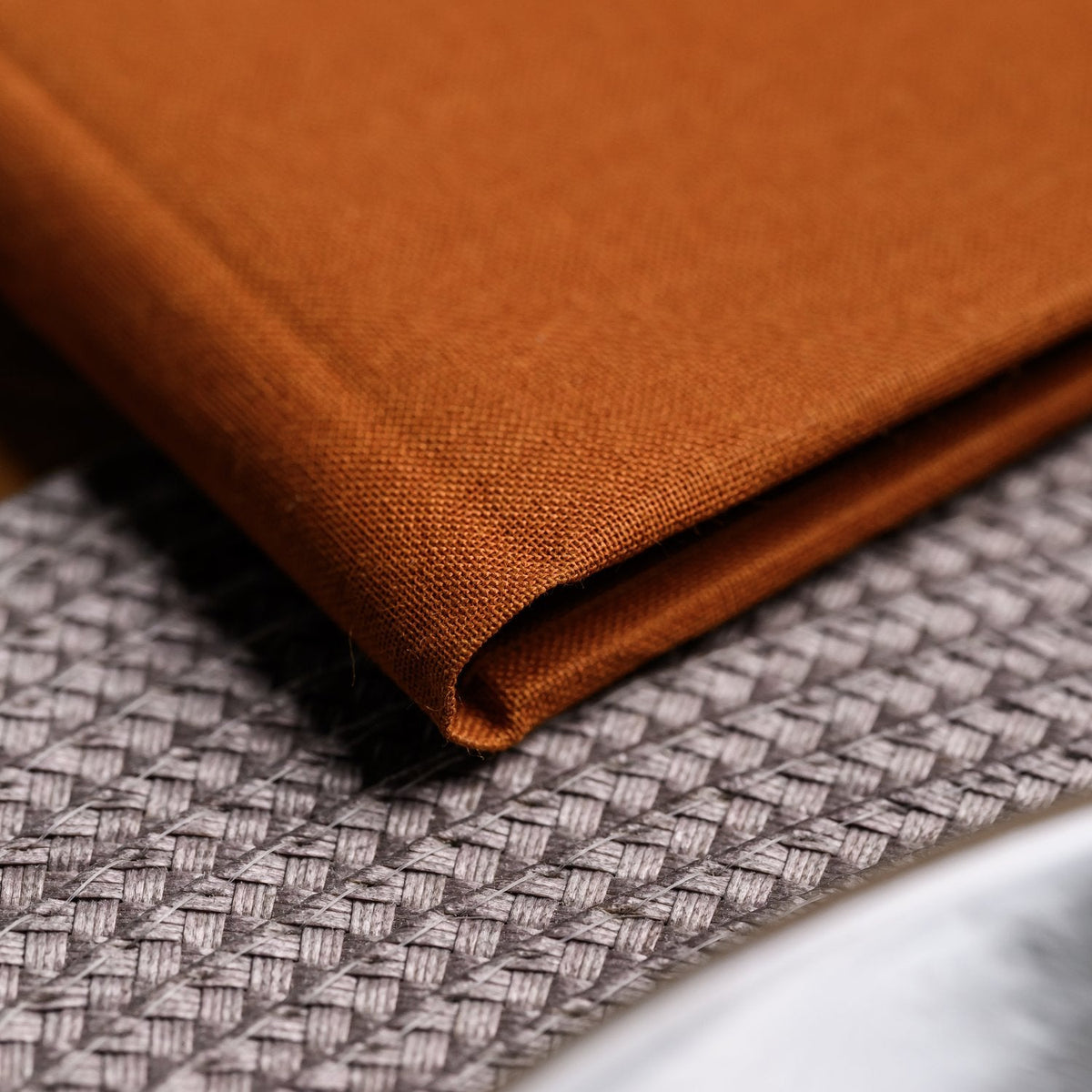 Close-up of a closed rust-orange textile folder corner beside a plate of food and a fork on a woven mat