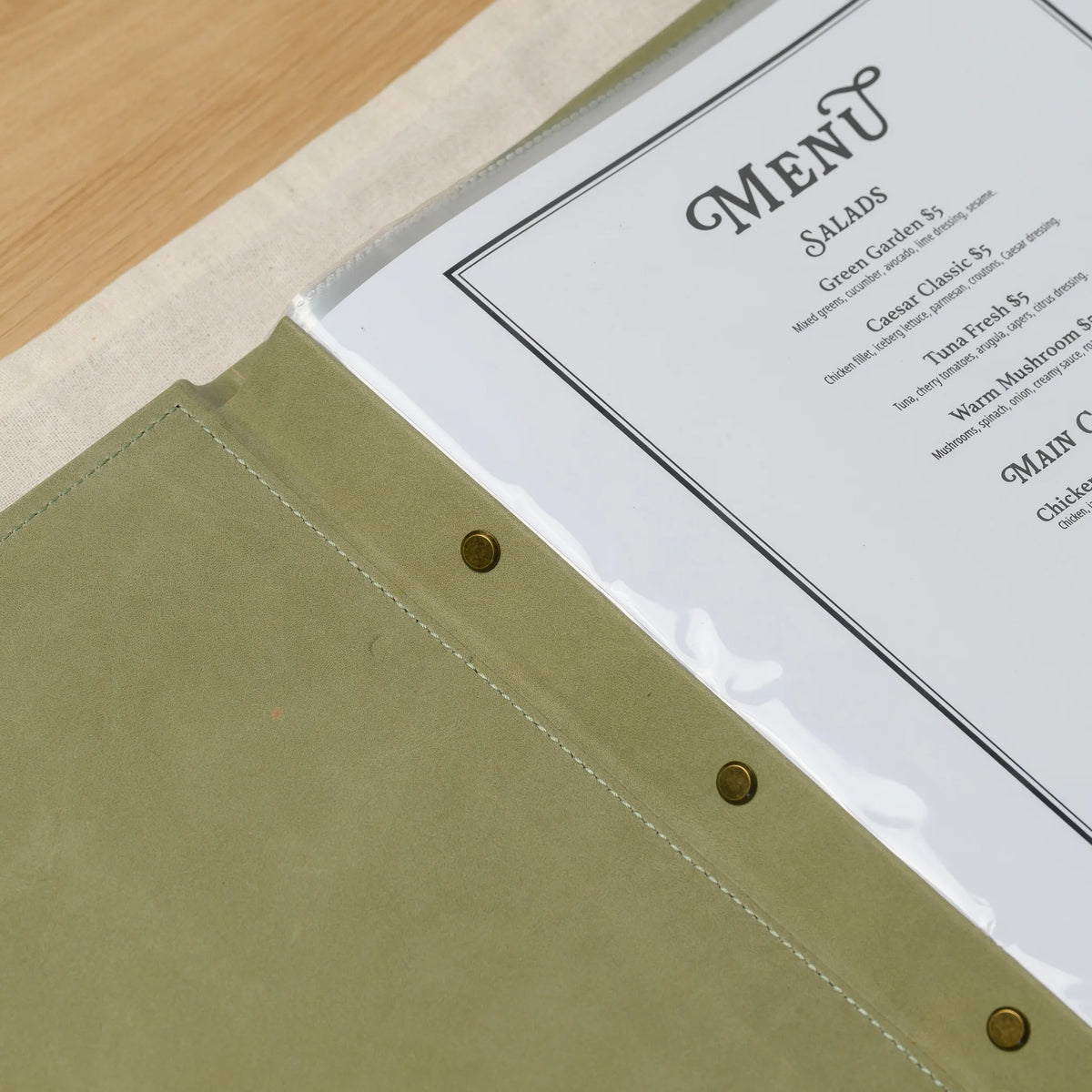 Close-up of three brass screws on the leather plank of an olive menu cover next to a printed menu in a clear sleeve