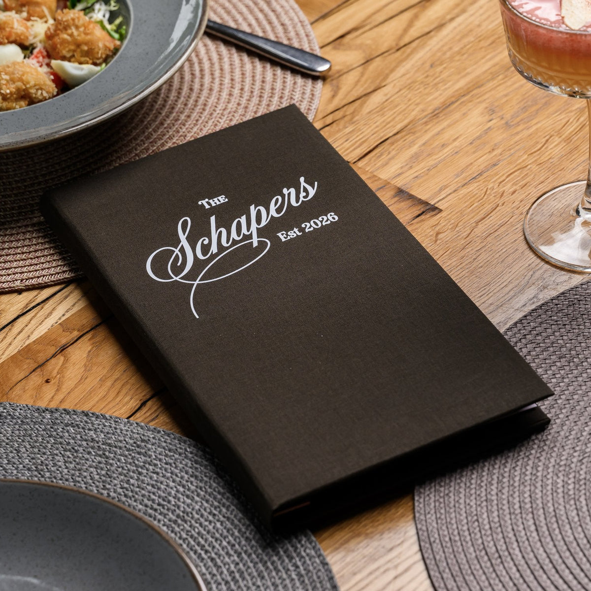 Small closed espresso-brown textile menu folder standing upright beside a wine glass on a woven placemat
