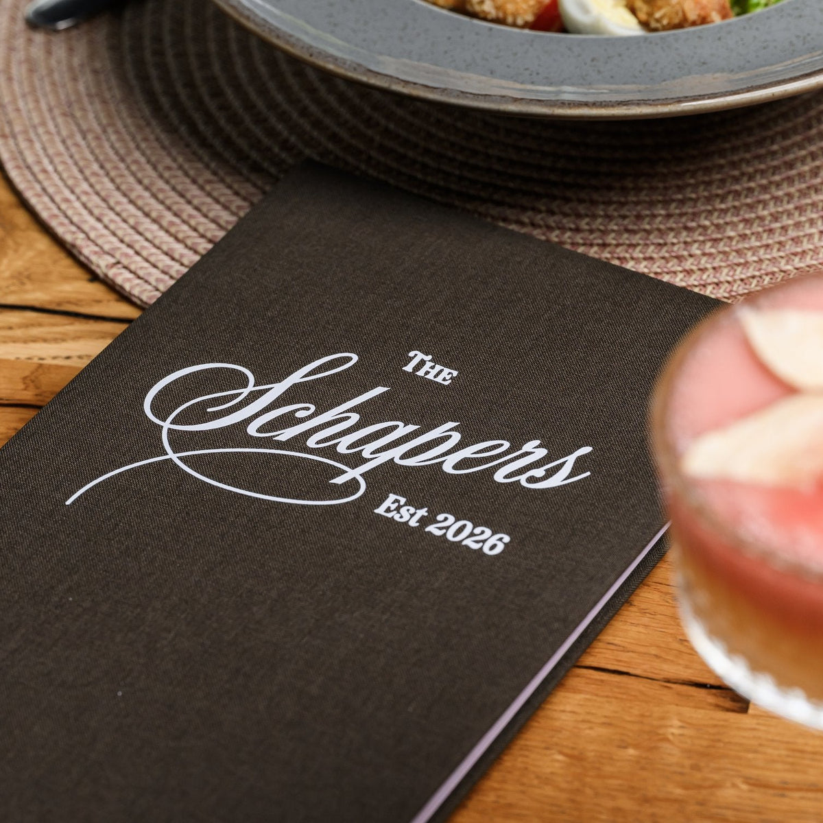Closed espresso-brown textile menu folder standing upright with a cursive logo beside a pink drink blurred