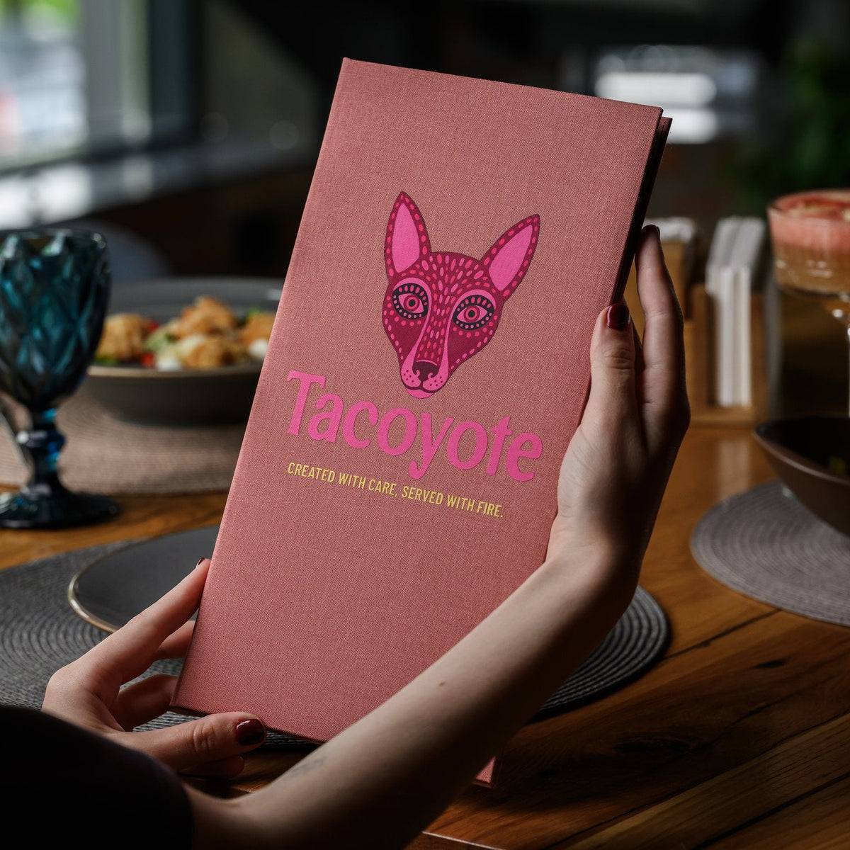 Hand holds a closed coral-pink textile menu folder with a fox graphic against a blurred cafe interior