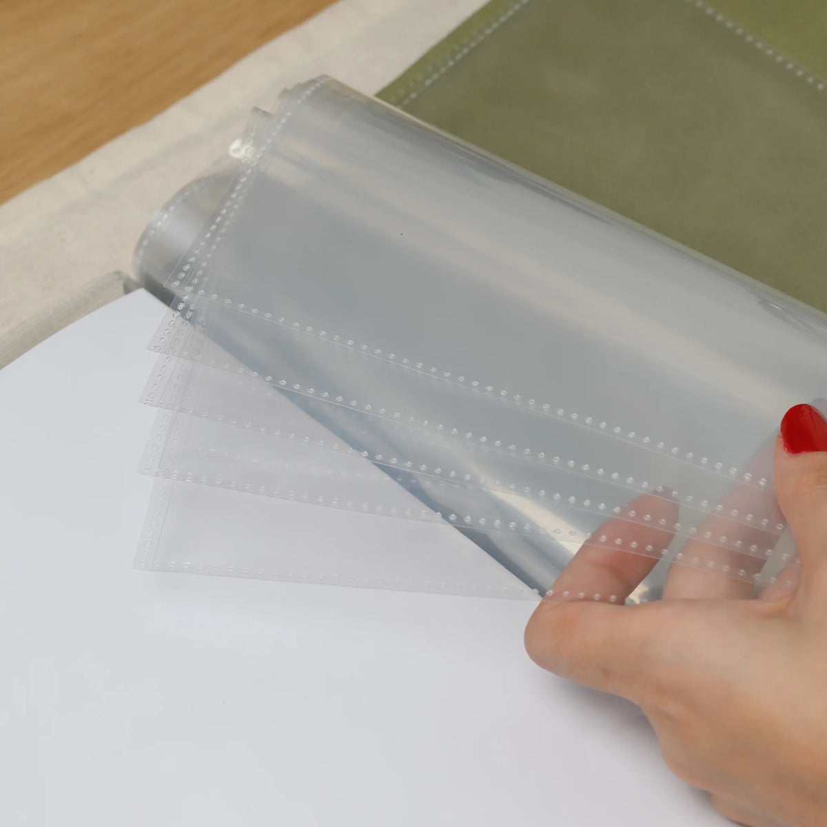 Hand fanning a stack of clear plastic menu sleeves with heat-sealed dotted seams above an olive faux leather cover