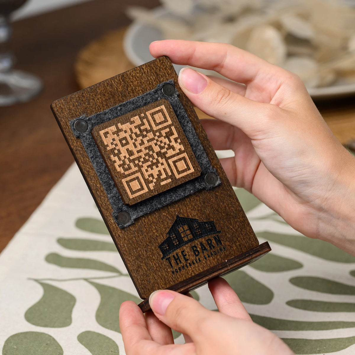A hand holding a wooden QR code table stand with a felt-framed engraved panel above a set dinner table