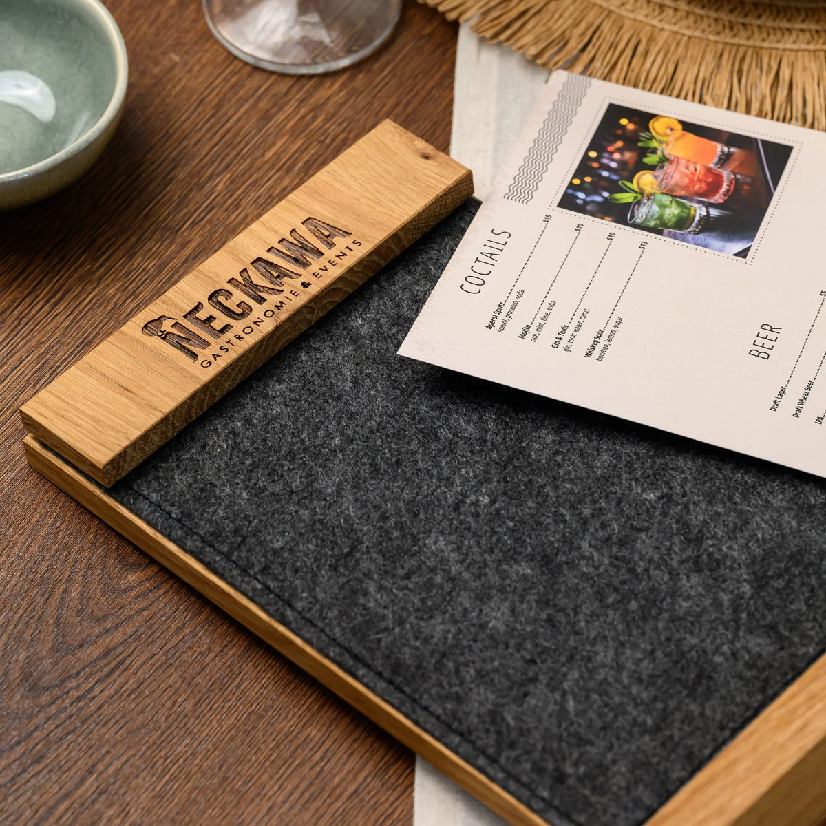 A freestanding table tent with an engraved oak bar tilted open, showing a printed cocktail menu page