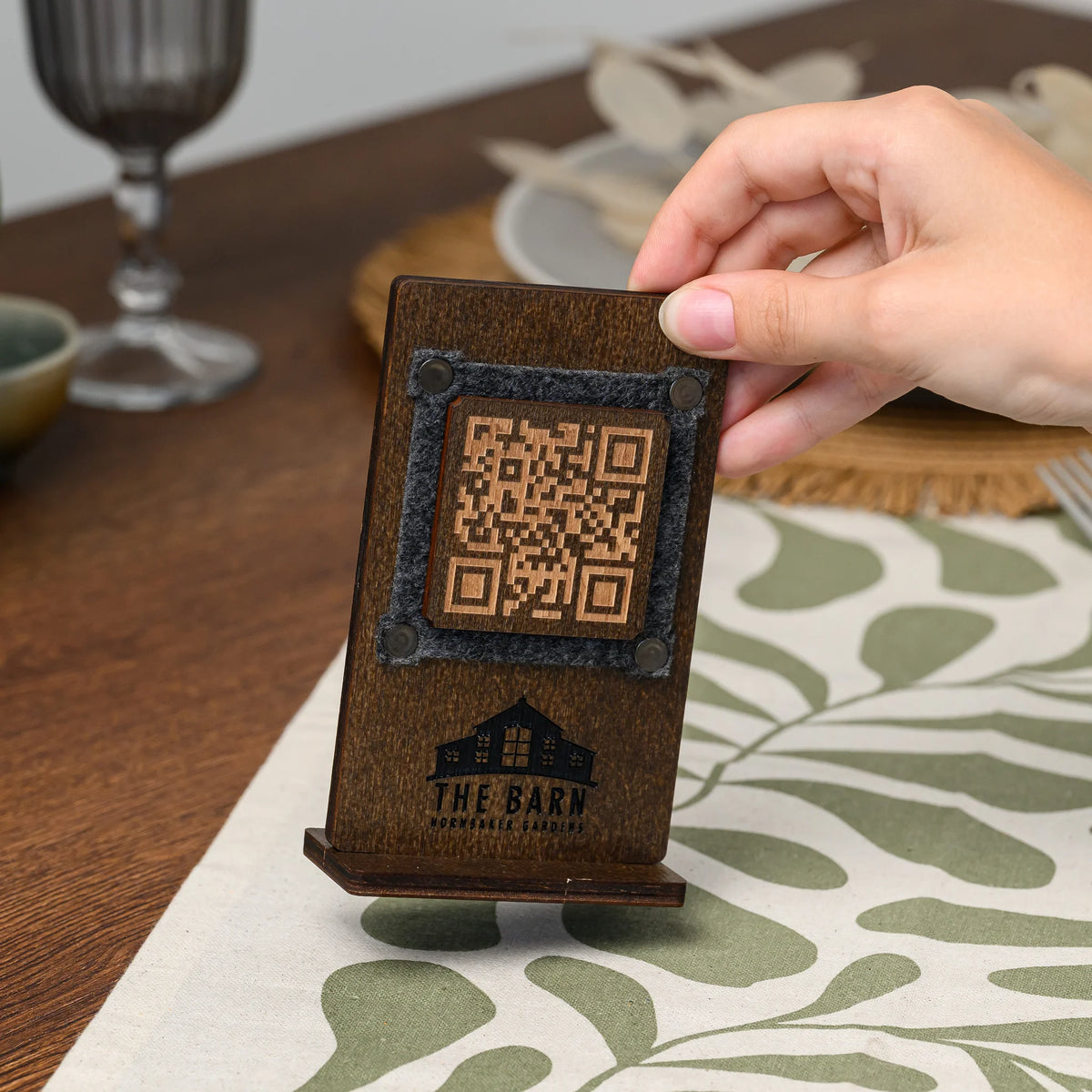 A hand holding a wooden QR code table stand near a plate, showing the felt-framed engraved panel closely