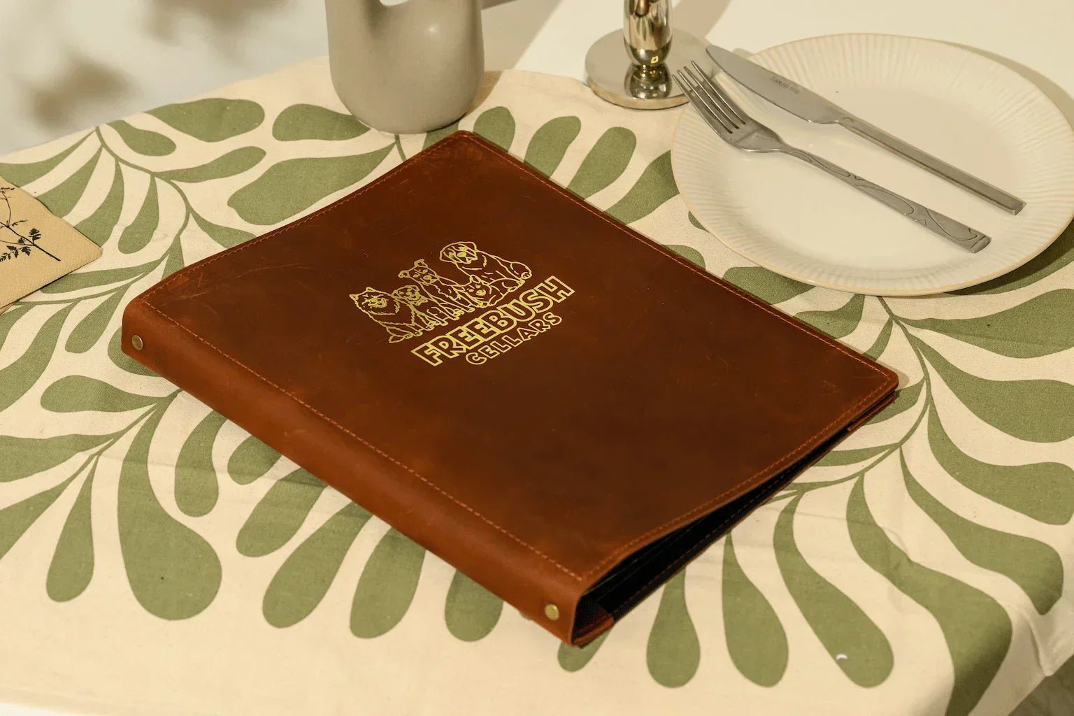 Handcrafted brown leather menu folder with custom gold foil branding and professional bolt binding, shown in a premium table setting