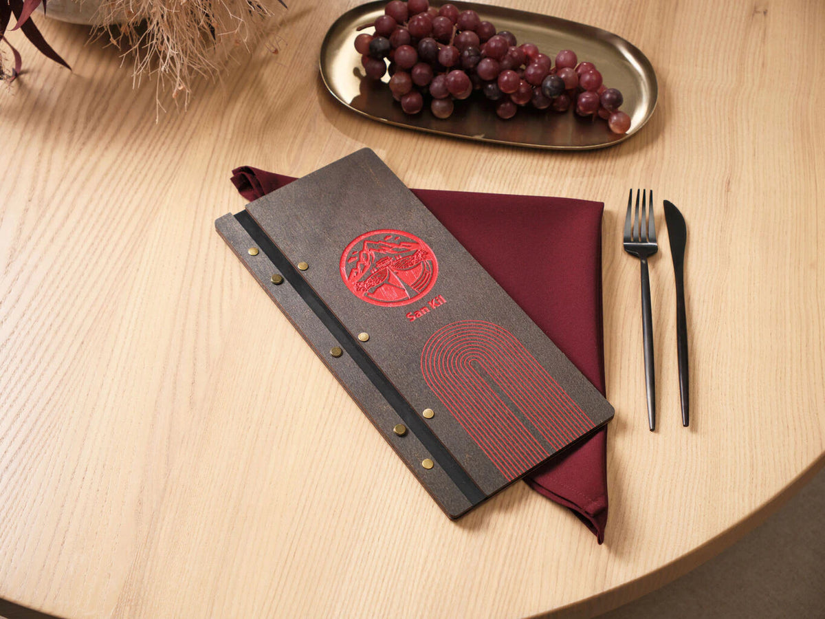 Dark wenge plywood slim menu folder with arch engraving and red round logo on wooden table, burgundy napkin beside