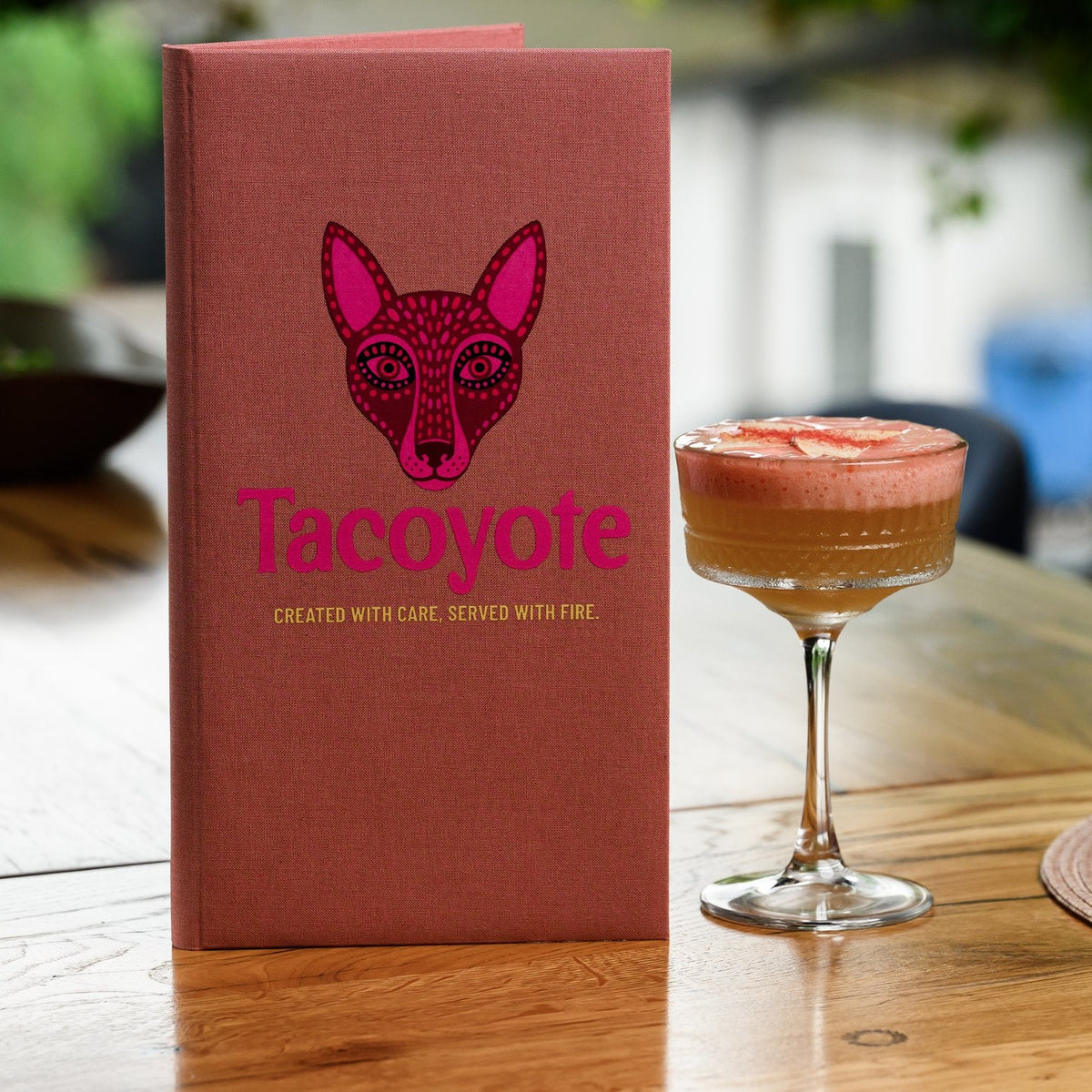Coral-pink textile menu folder with a bold fox graphic standing tall on a wooden table beside a cocktail