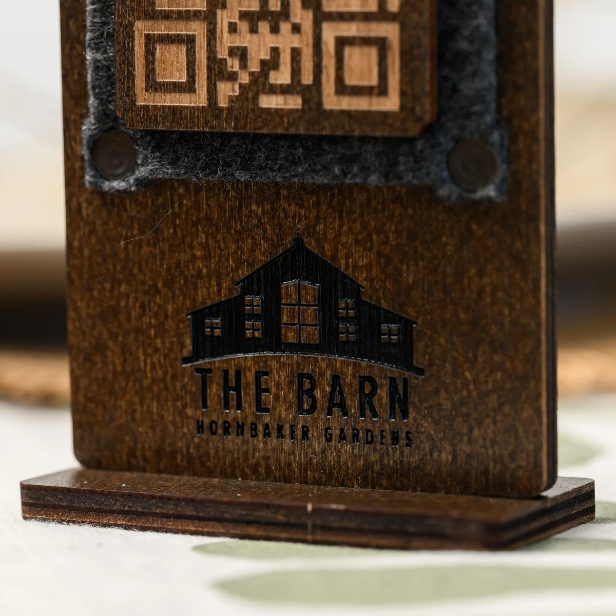 Close-up of an engraved logo beneath a felt-framed QR code panel on a wooden table stand, front view