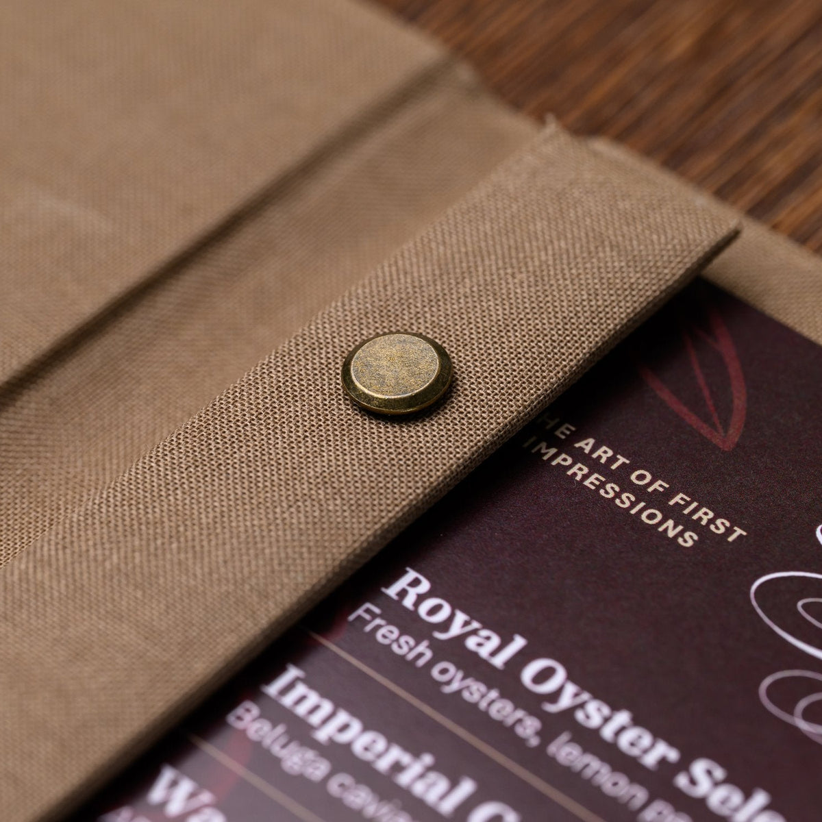 Closed taupe textile menu folder with a snap fastener and a dark inner menu page peeking out on the table