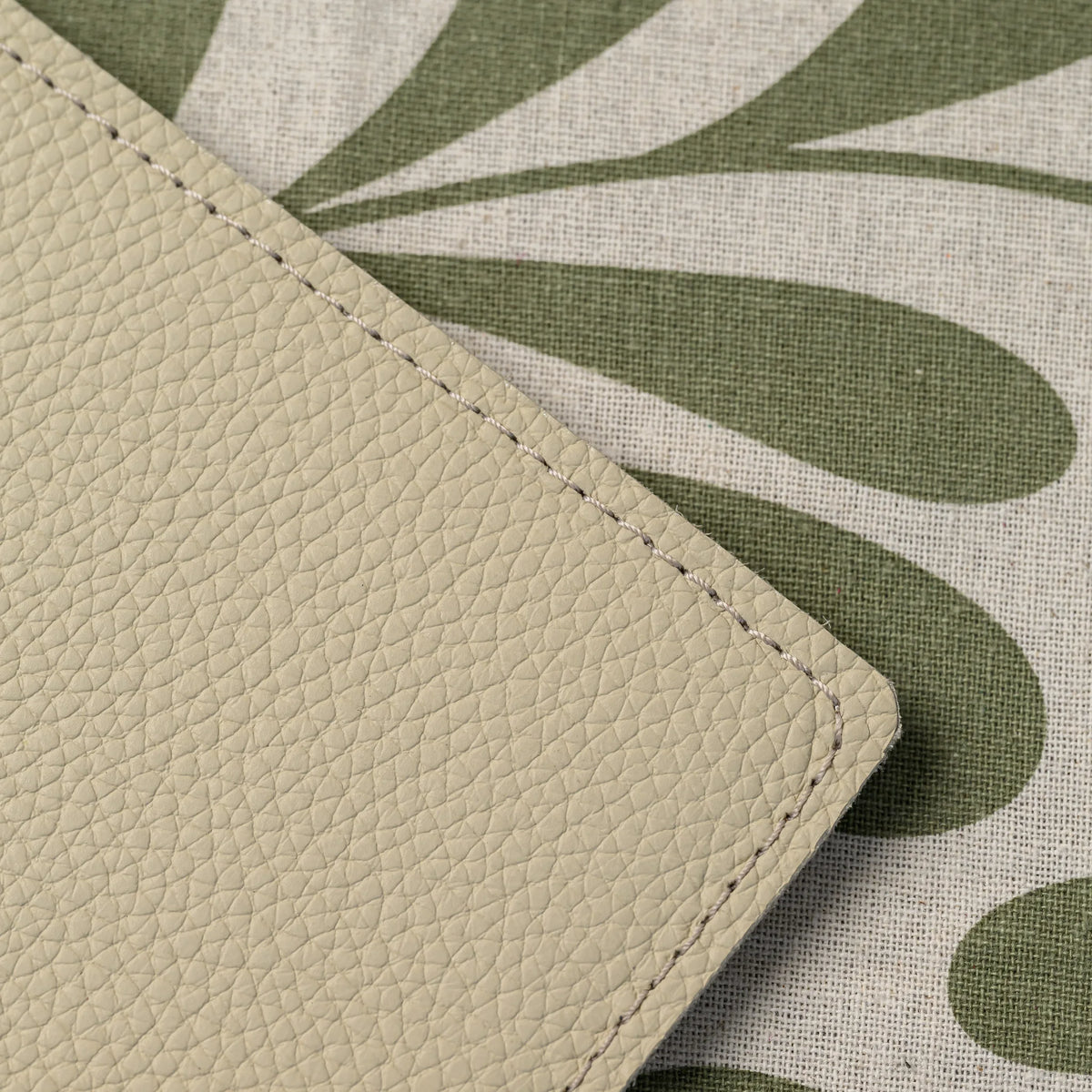 Close-up of beige pebble-grain leather cover corner with tonal stitching lying on a leaf-print linen table runner