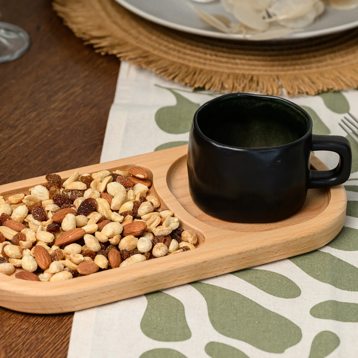 Black ceramic mug placed in one oval compartment beside mixed nuts in the other, on the beech wood board