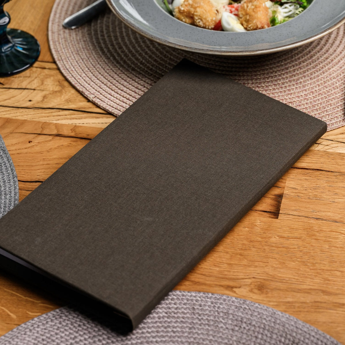 Closed espresso-brown textile menu folder lying flat on a wooden table beside a grey woven table placemat