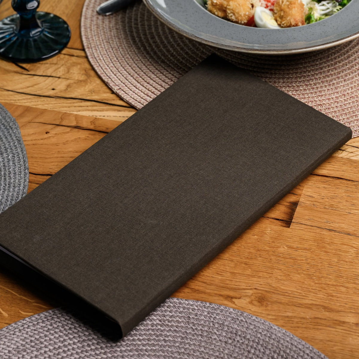 Closed espresso-brown textile menu folder lying flat on a wooden table beside a woven placemat, angled view